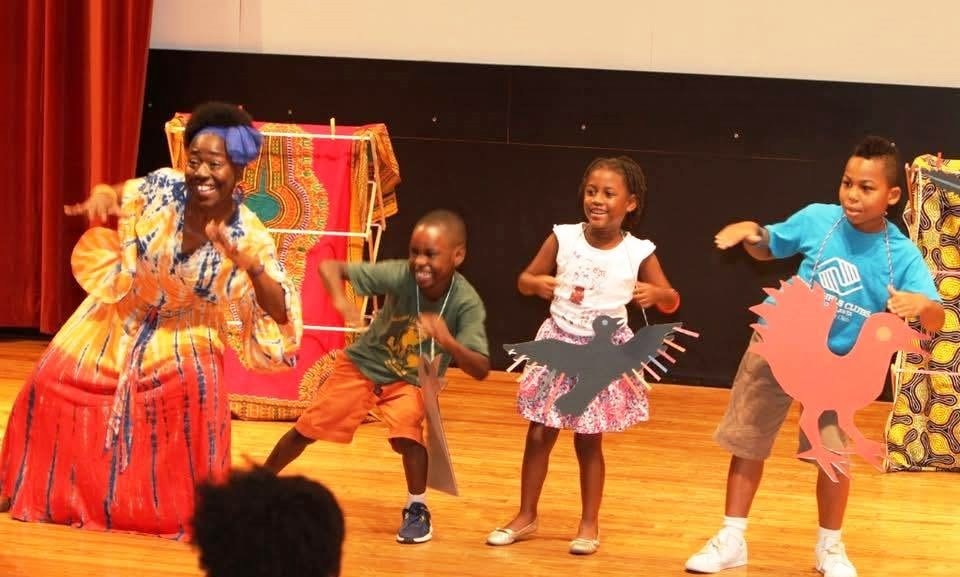 Mama Koku dancing on stage with children holding bird and rooster cutouts